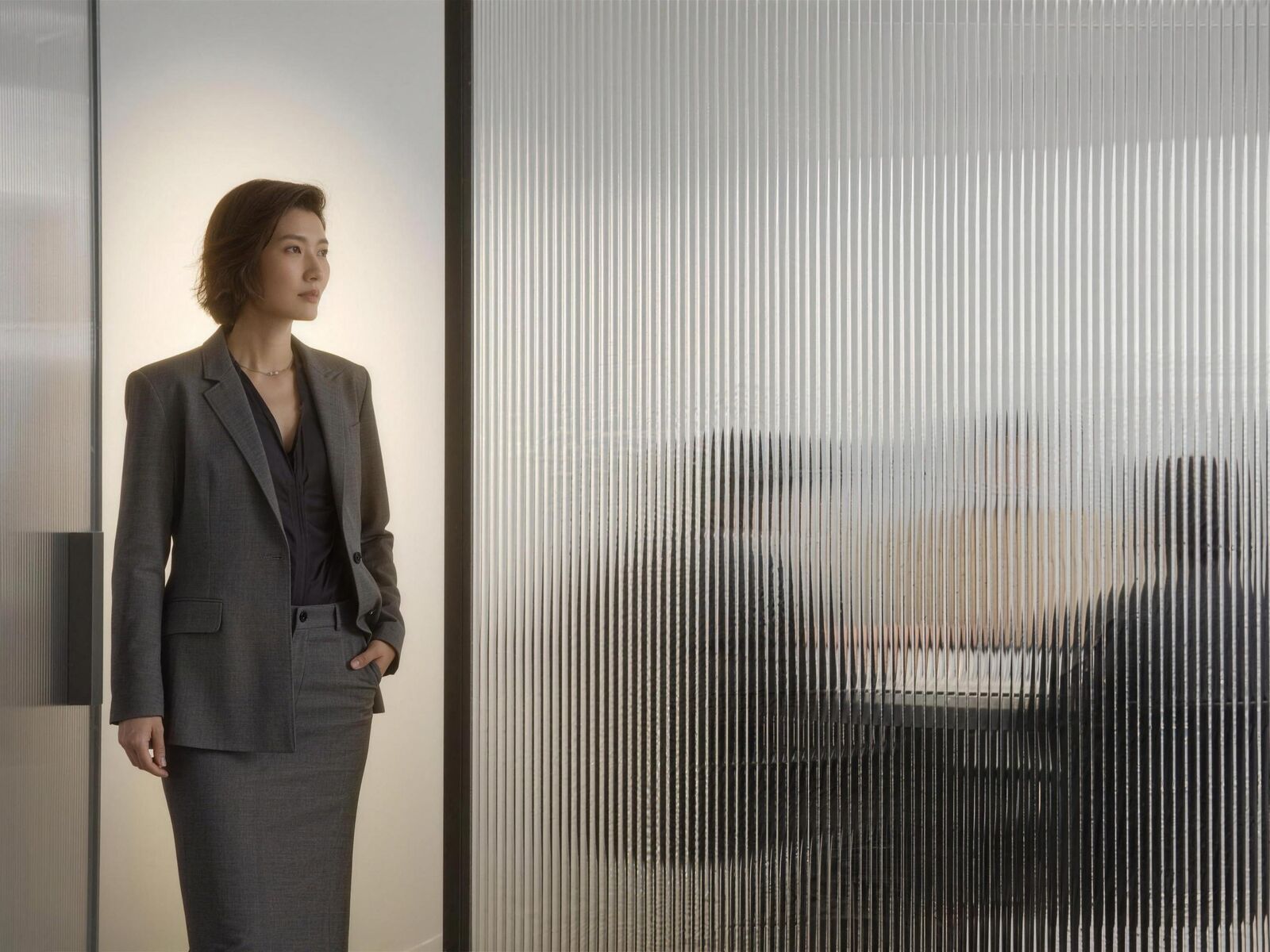 A woman in a gray suit stands by a ribbed glass wall; behind it, a meeting in soft focus